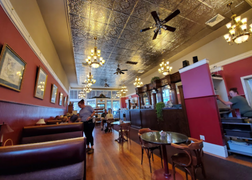 A cozy restaurant interior with vintage decor, chandeliers, and patrons dining at tables. Staff are serving and cleaning.