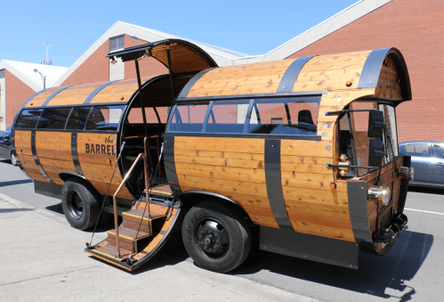 A wooden barrel-shaped food truck with large windows and a staircase, parked on a city street.