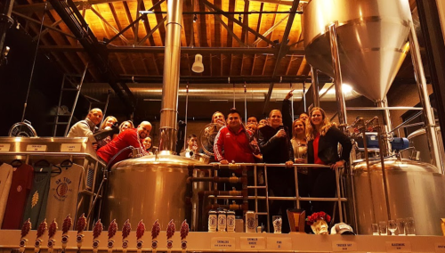A group of people smiling and posing together in a brewery, surrounded by brewing equipment and tanks.