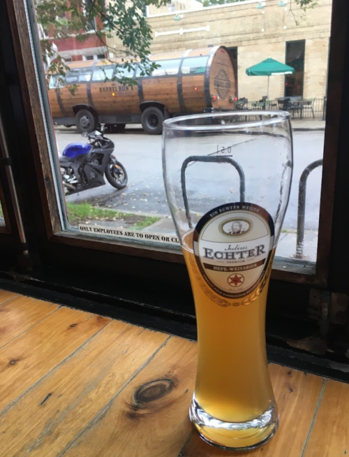 A glass of beer sits on a wooden table, with a view of a street and a parked motorcycle outside the window.