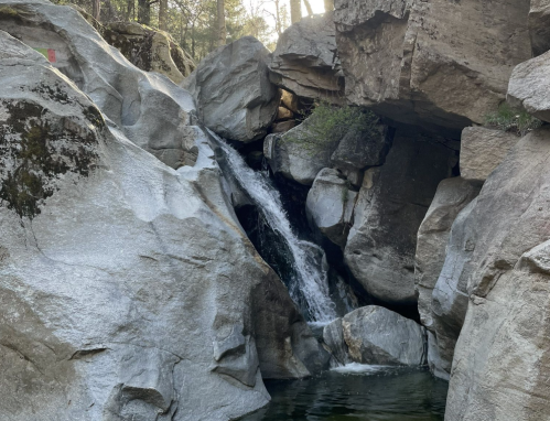 A small waterfall cascades over rocks into a serene pool, surrounded by rugged stone formations and trees.