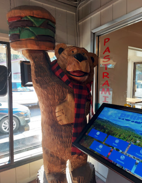 A wooden bear statue holding a burger, wearing a red and black checkered scarf, inside a restaurant.