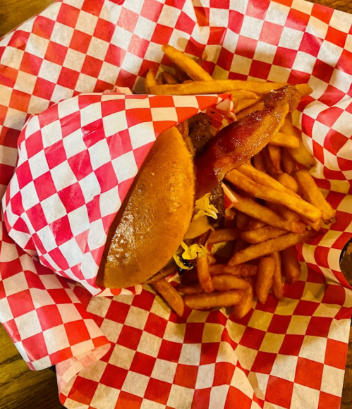 A wrapped sandwich with bacon and toppings sits beside a pile of crispy fries, all on red and white checkered paper.