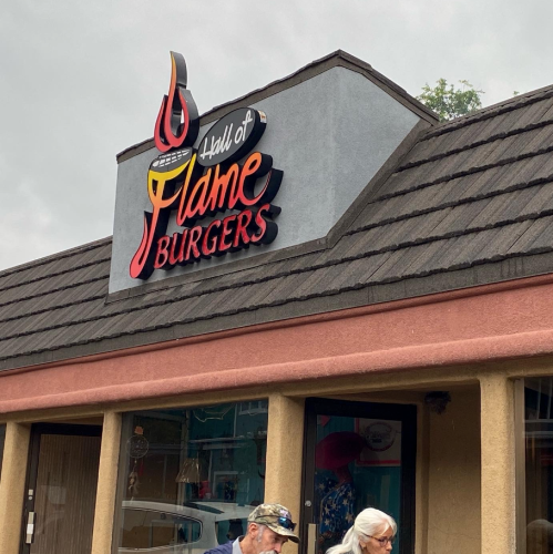 Sign for "Hall of Flame Burgers" on a building with a gray roof and pink walls, featuring two people walking by.
