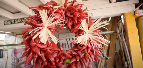 A vibrant wreath made of red chili peppers, adorned with straw accents, hanging in a rustic setting.