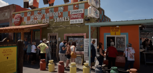 A busy restaurant exterior with colorful signage, people waiting in line, and a vibrant atmosphere.