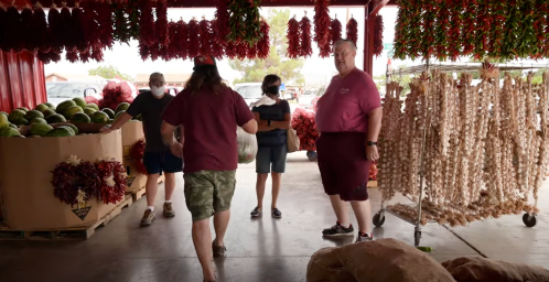 A group of people in masks at a market, surrounded by watermelons and hanging decorations.