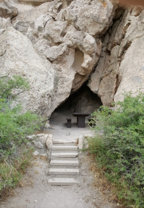 A rocky cave entrance with a small table and bench, surrounded by greenery and stone steps leading inside.