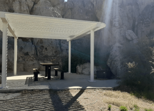 A shaded picnic area with a table and benches, surrounded by rocky terrain and sparse vegetation.
