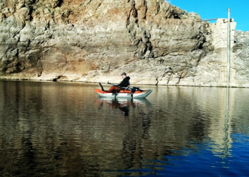 A person fishing from a small kayak on calm water, surrounded by rocky cliffs and clear blue skies.