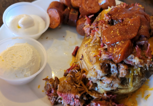 A plate featuring a loaded baked potato topped with meat and sauce, alongside sliced sausage and two cups of sour cream.
