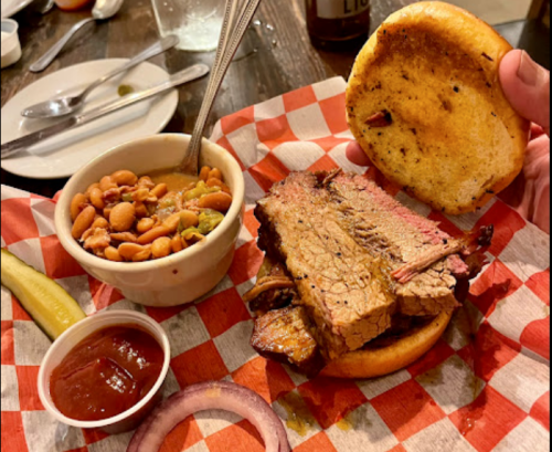 A hand holds a sandwich with brisket on a bun, accompanied by baked beans, pickles, onion, and a side of ketchup.