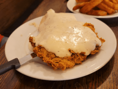 A plate of fried chicken smothered in creamy gravy, with a side of crispy fries in the background.