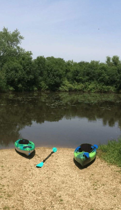 Two green kayaks and a paddle sit on a gravel shore by a calm, reflective river surrounded by lush greenery.