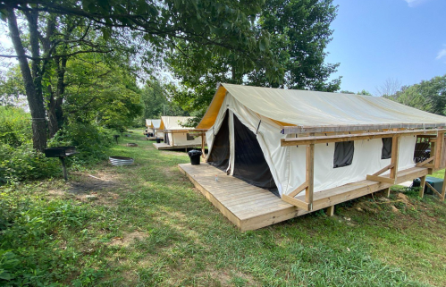 Two canvas tents on wooden platforms in a grassy area, surrounded by trees and blue sky.