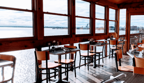 Bright restaurant interior with wooden walls, tables, and chairs, overlooking a scenic waterfront view through large windows.
