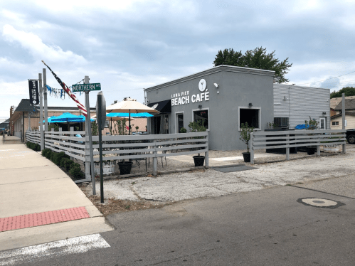 A gray cafe with outdoor seating, located at a street corner, under a cloudy sky. Flags and umbrellas are visible.