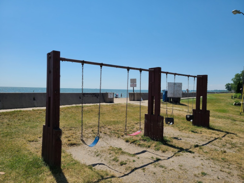 A playground with swings on a sunny day, overlooking a calm lake and clear blue sky.