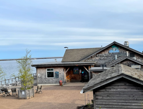 A rustic café named "Angry Trout Café" by the water, with wooden buildings and a calm sky in the background.