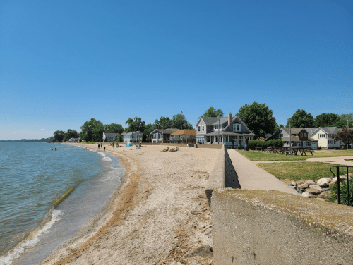 A sandy beach with calm water, lined by houses and trees under a clear blue sky. People enjoy the shoreline.