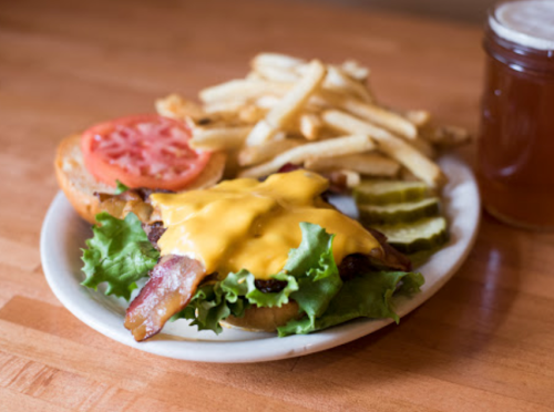 A cheeseburger with bacon, lettuce, tomato, and pickles, served with fries and a drink in a glass.