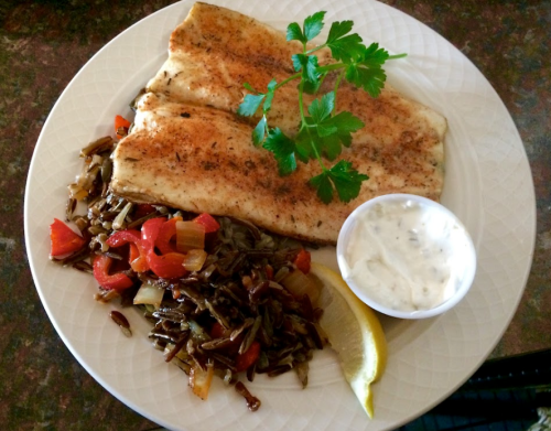 A plate with seasoned fish fillets, sautéed vegetables, a lemon wedge, and a side of dipping sauce, garnished with parsley.