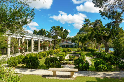 A serene garden with manicured hedges, stone benches, and a pergola under a bright blue sky with fluffy clouds.