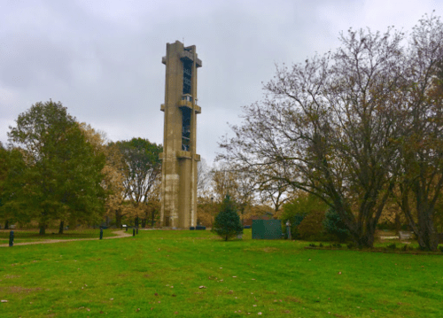 A tall concrete tower stands in a park surrounded by trees and green grass under a cloudy sky.