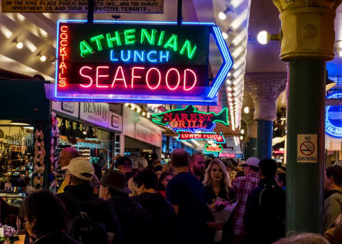 A bustling market scene with neon signs for "Athenian Lunch" and "Seafood," surrounded by a crowd of people.