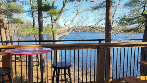 A scenic view of a lake surrounded by trees, seen from a wooden deck with a table and chairs.