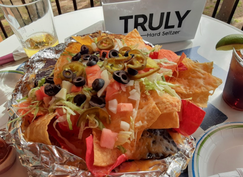 A large plate of nachos topped with lettuce, tomatoes, jalapeños, and olives, served with drinks in the background.
