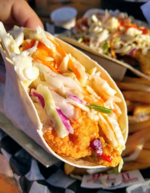 A close-up of a fish taco filled with crispy fish, coleslaw, and sauce, with fries in the background.