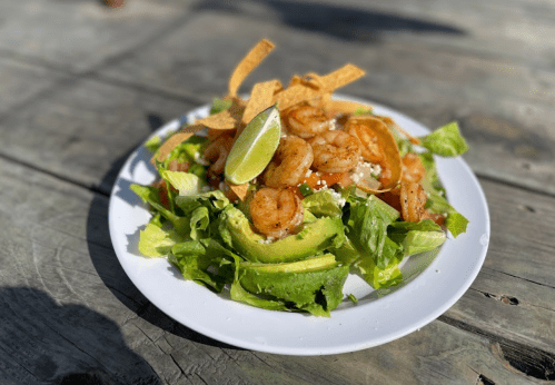 A vibrant shrimp salad with avocado, lettuce, lime, and tortilla strips on a white plate, set on a wooden table.