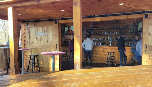 A rustic bar with wooden walls, shelves of bottles, and patrons sitting at the counter and tables.