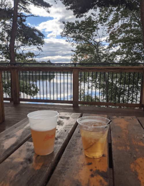 Two drinks on a wooden table overlooking a serene lake and trees under a cloudy sky.
