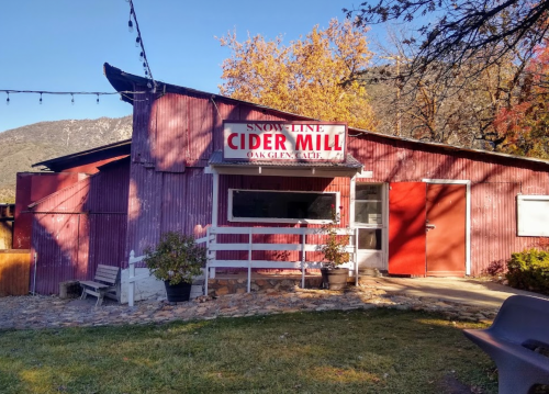 A rustic cider mill with a red exterior, sign reading "Snow-Line Cider Mill," surrounded by trees and mountains.