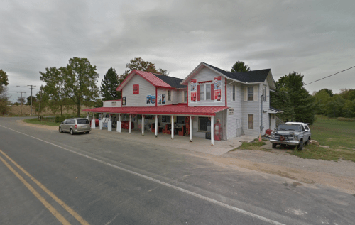 A two-story building with a red roof, featuring a storefront and parked cars along a rural road.