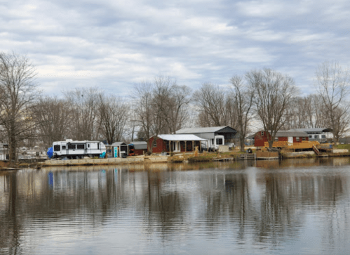 A peaceful lakeside scene featuring houses and an RV along the shore, with trees and cloudy skies in the background.