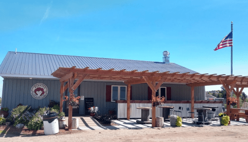 A rustic building with a metal roof, wooden pergola, and an American flag, surrounded by plants and outdoor seating.