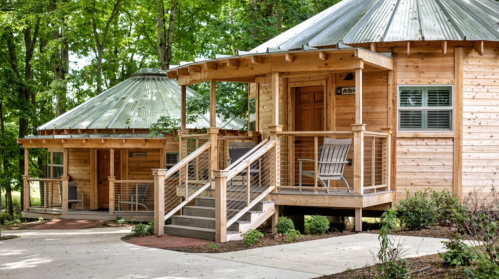 Two wooden cabins with metal roofs, surrounded by trees, featuring porches and steps leading to the entrance.