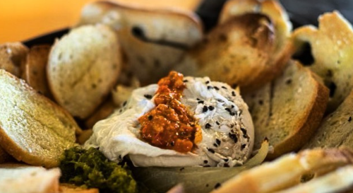 A bowl of toasted bread slices surrounding a creamy dip topped with red sauce and sprinkled with black seeds.