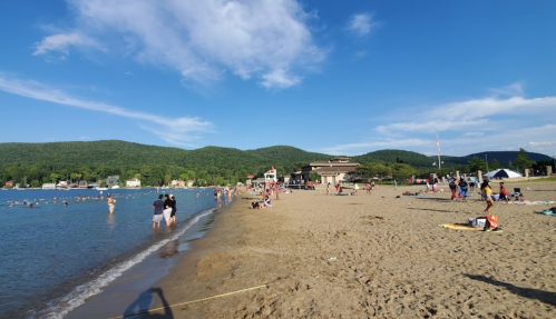 A sandy beach with people swimming and relaxing, surrounded by green hills and a clear blue sky.