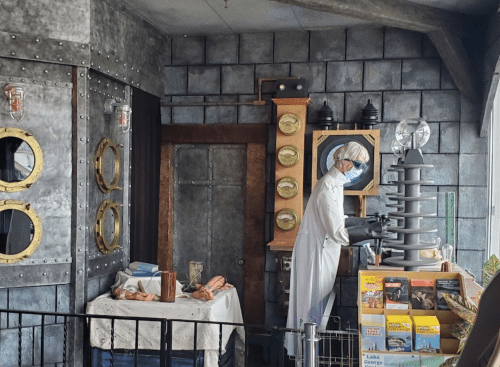 A scientist in a lab coat works with equipment in a steampunk-style room filled with gadgets and books.