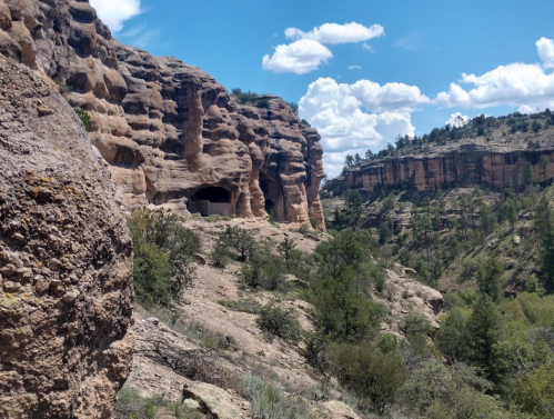 Rocky cliffs and caves under a bright blue sky with scattered clouds, surrounded by greenery in a rugged landscape.