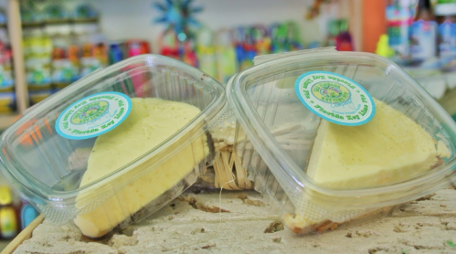 Two slices of cheese in clear plastic containers, displayed on a rustic surface with colorful items in the background.