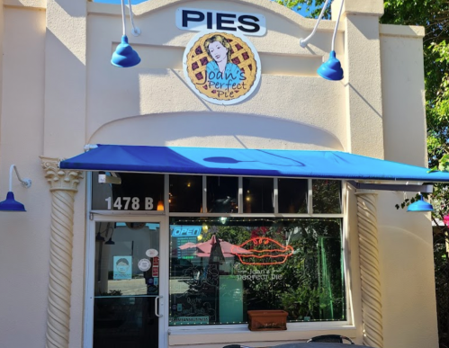 Exterior of a pie shop named "Joan's Perfect Pie," featuring a blue awning and a sign with a woman's face and pie graphic.