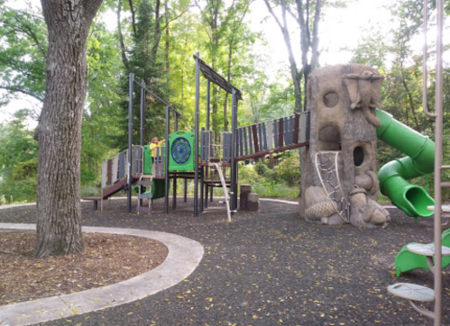 A playground with a climbing structure, slide, and bridge surrounded by trees and a gravel path.
