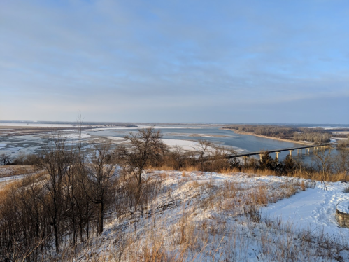A scenic view of a river winding through a snowy landscape, with trees and a bridge in the distance under a cloudy sky.