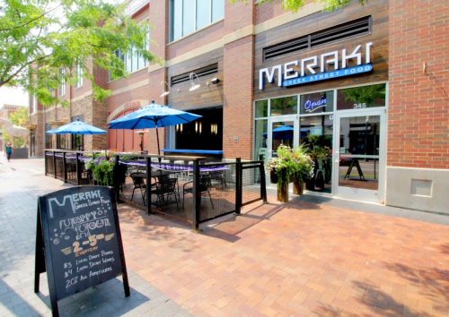 Exterior of Meraki Greek Street Food with outdoor seating, umbrellas, and a chalkboard menu displaying specials.
