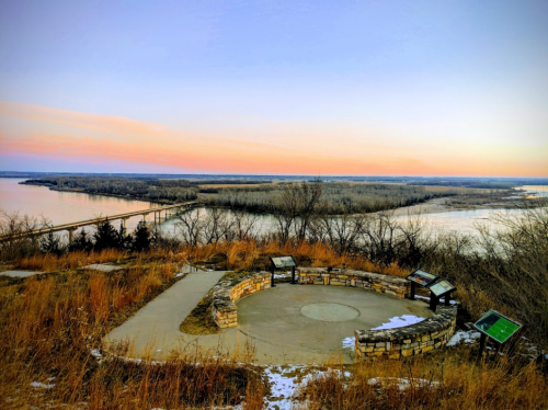 Scenic view from a lookout point over a river, surrounded by trees and grass, with a colorful sunset sky.